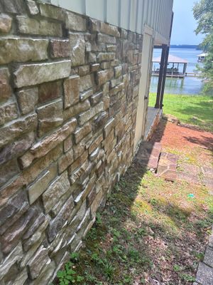 Exterior view of a stone foundation wall along a waterfront property with overgrown grass and exposed soil at the base,