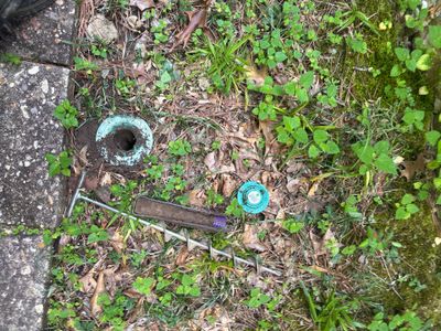 An exterior catch basin or drain box with a blue marker is visible in a grassy, weedy yard area during installation or
