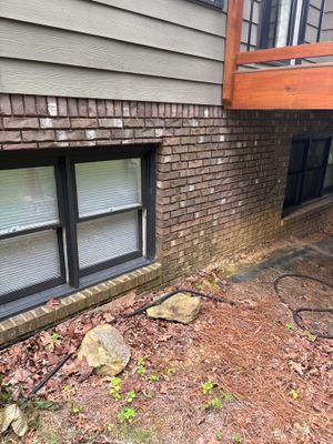 Exterior foundation area showing brick veneer, siding, basement windows, and bare ground with minimal vegetation,