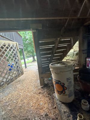 An exterior view of a covered carport or storage area with gravel ground, showing the structure's underside and support