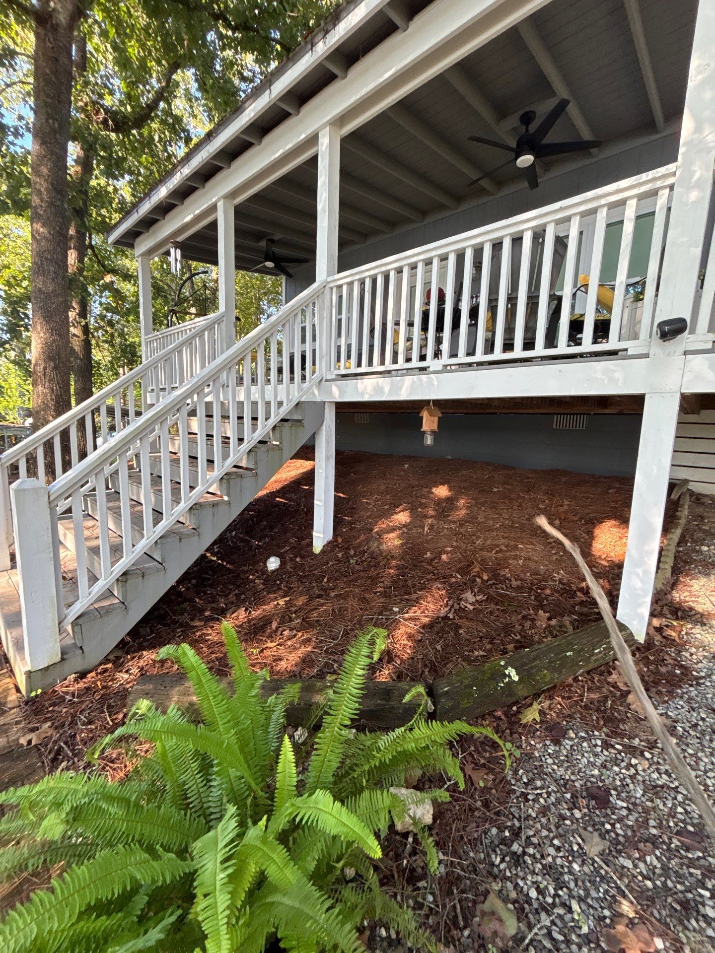 A residential house with a covered deck/porch, white railings, and landscaped ground beneath, showing the exterior
