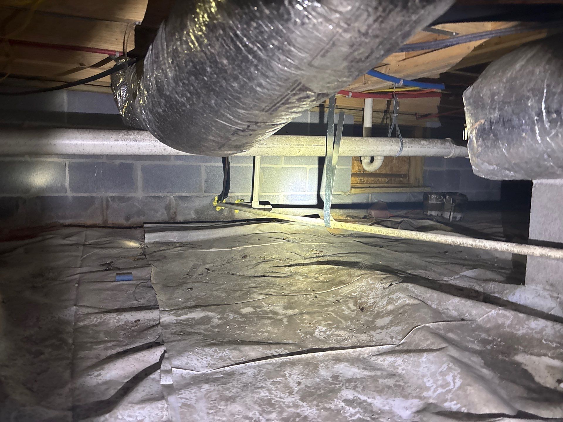 A crawl space interior showing plastic vapor barrier installation and drainage systems in progress, with support posts,