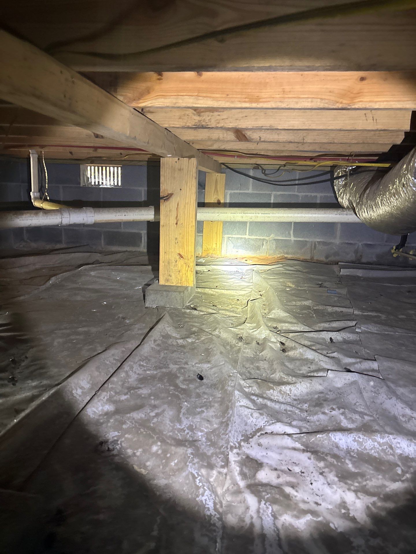 A crawlspace during active encapsulation work showing vapor barrier installation on the floor, wooden support posts,